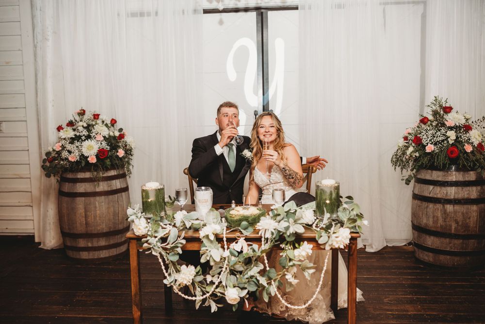 A newlywed couple sits at their decorated sweetheart table with rustic wine barrel and floral arrangements.