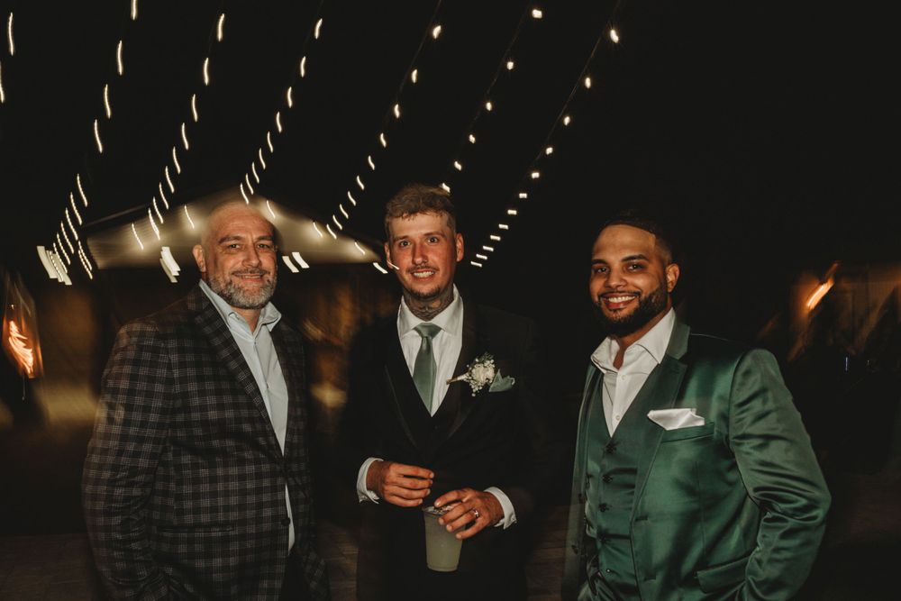 Three wedding guests in formal attire pose together under string lights at night.