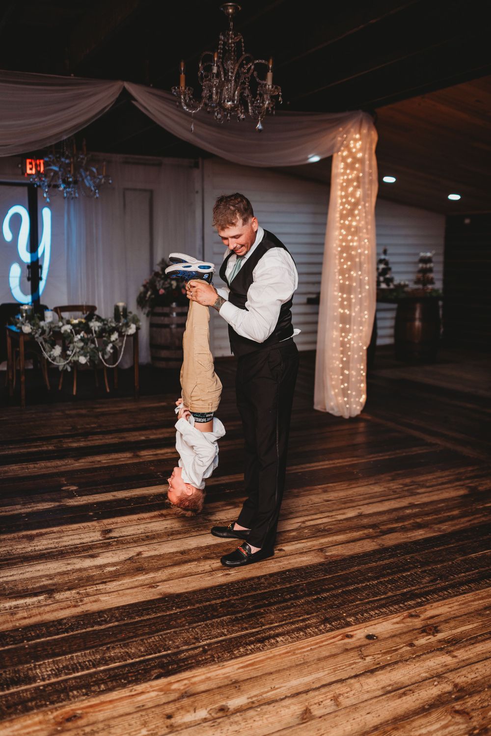 An adult dances with a child at a wedding reception under elegant chandeliers and draping.