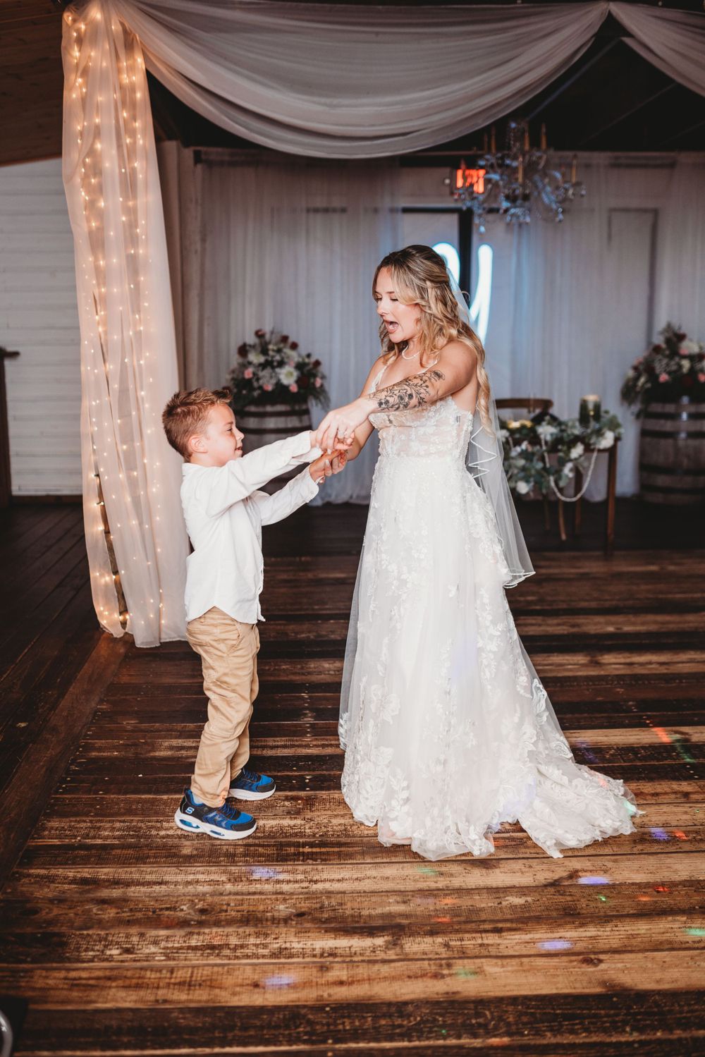 A young child dances with someone wearing a white wedding gown on a wooden dance floor decorated with lights and drapes.