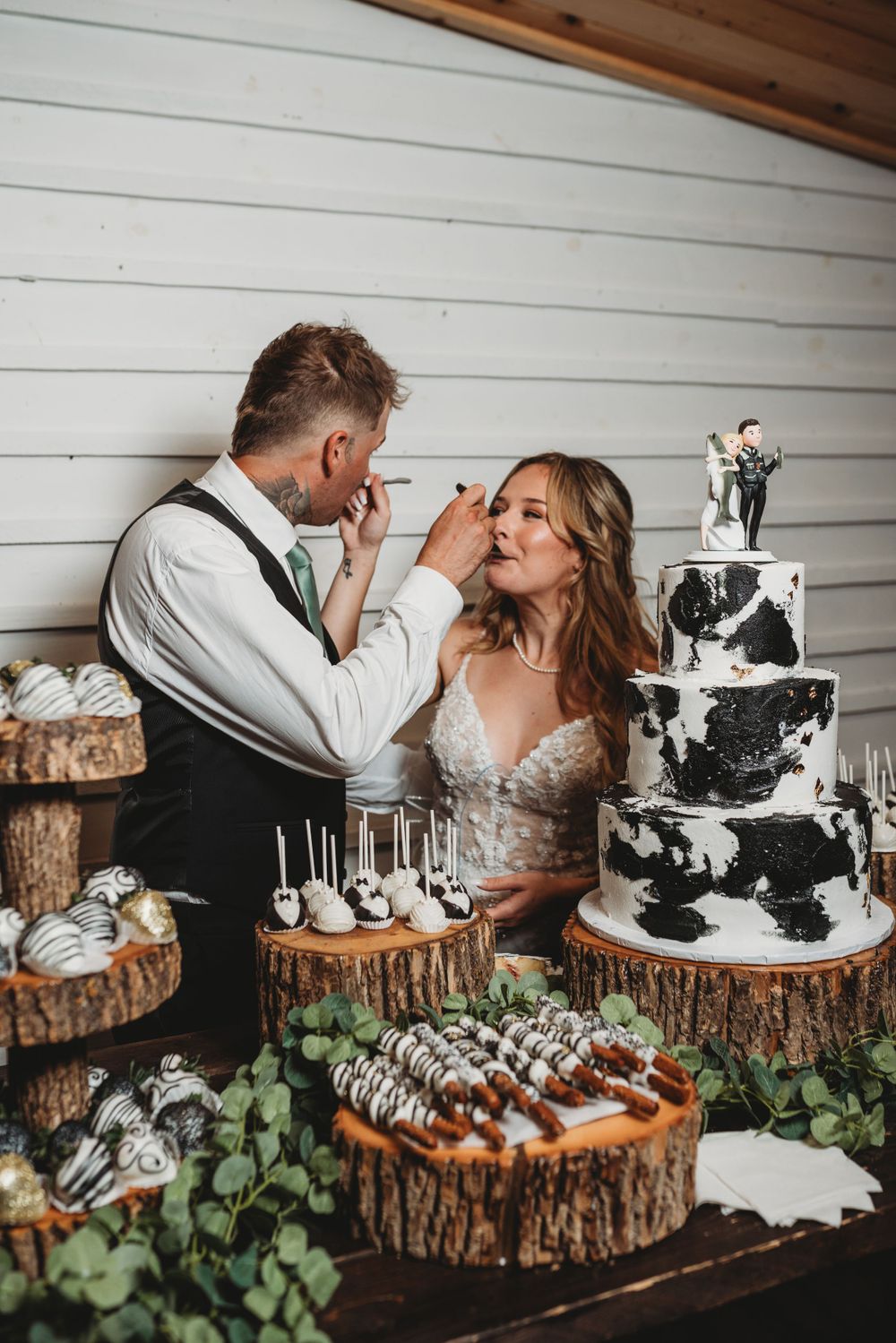 A couple feeds each other cake at their rustic-themed wedding reception with a black and white tiered cake.
