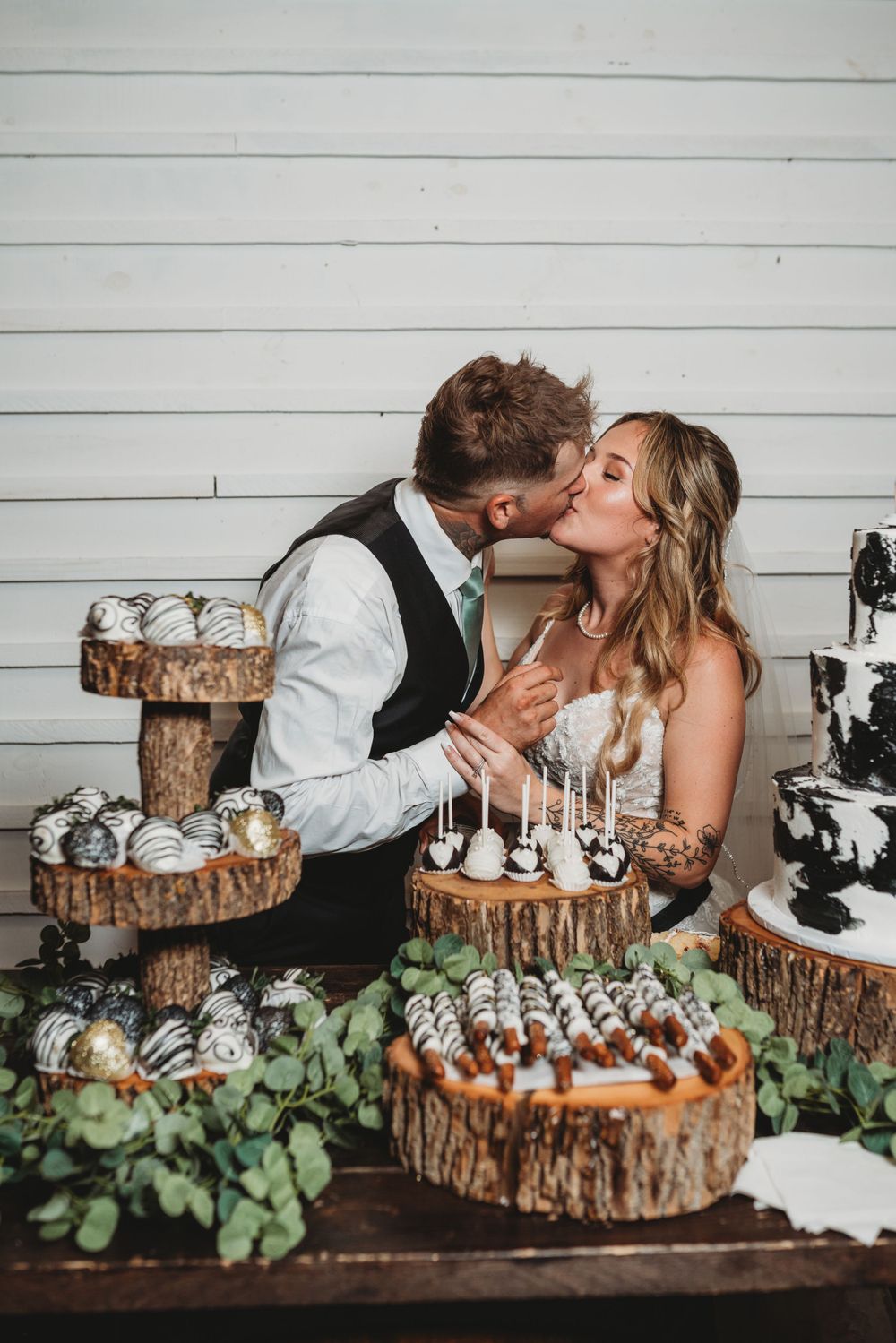 Newlyweds share a playful moment at their dessert table decorated with wood slice stands and greenery.