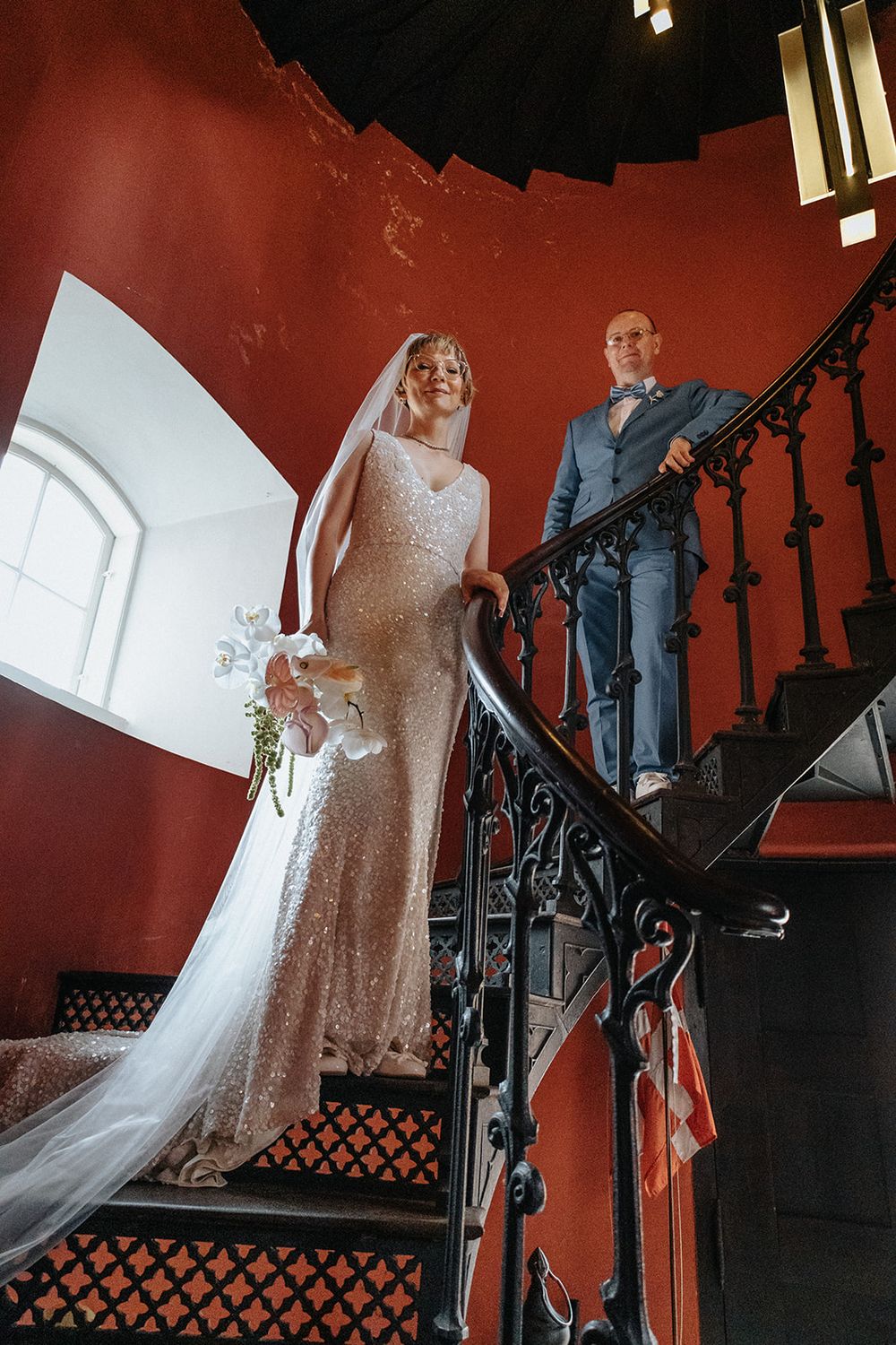 A wedding couple descends an elegant staircase with red walls and ornate black railings in a dramatic interior setting.