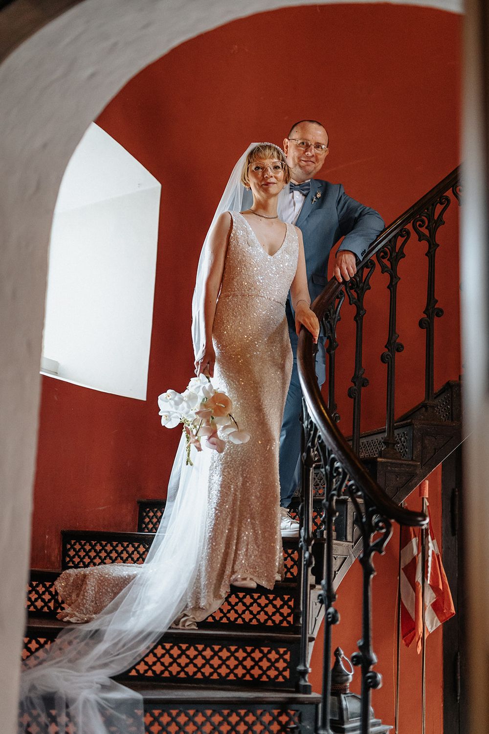 A couple poses on a red-walled staircase for wedding photos, with the bride's veil flowing elegantly behind them.