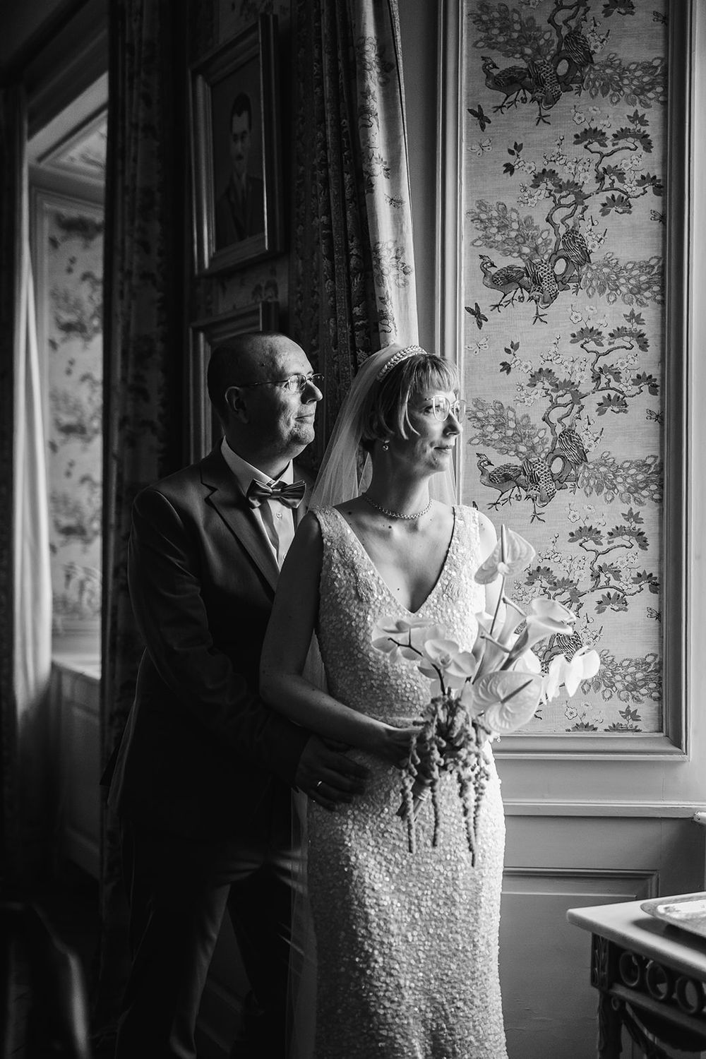A romantic black and white photo series showing a couple in formal attire by a window with floral wallpaper.