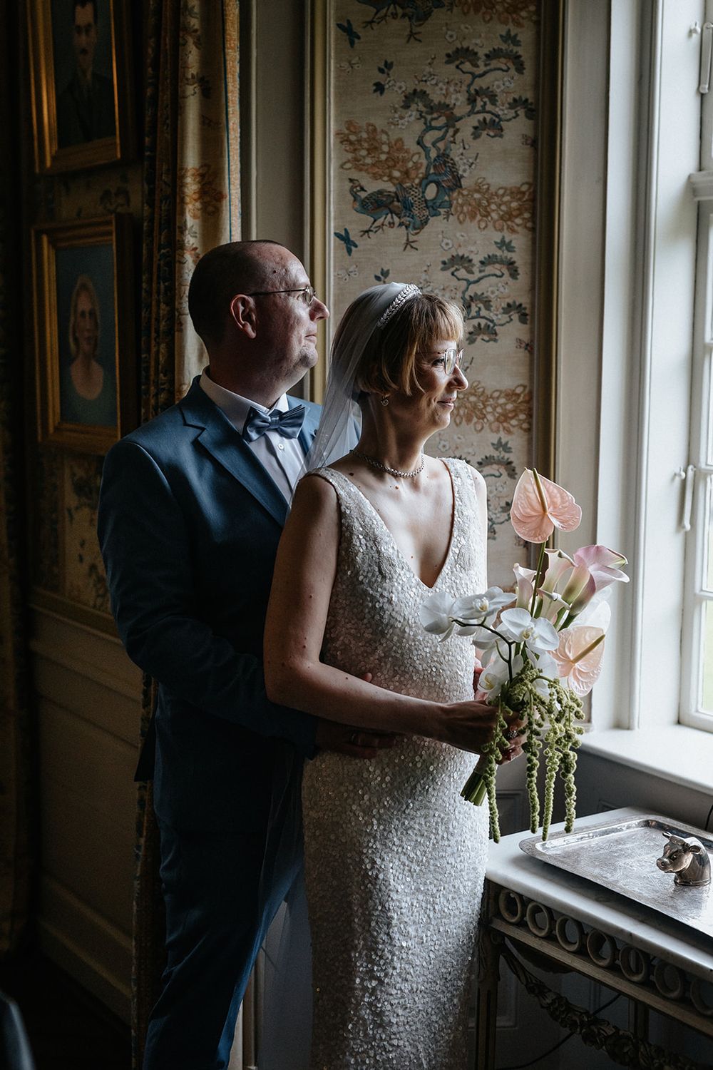 Couple in formal attire gazes through ornate windows in an elegant room with patterned wallpaper.