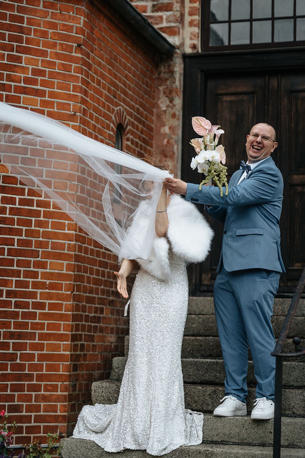 A couple shares a playful moment with the bride's veil on brick church steps during their wedding photoshoot.