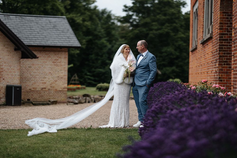 A bride and groom share a romantic moment together on a lawn with vibrant purple flowers in the background.