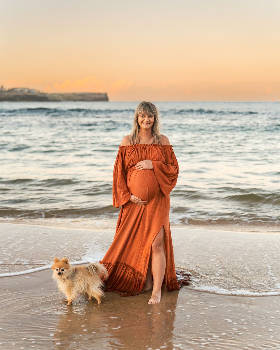 mom to be and her dog at sunset on a beach as gentle waves roll in around them during a pregnancy photoshoot in Maroubra Beach