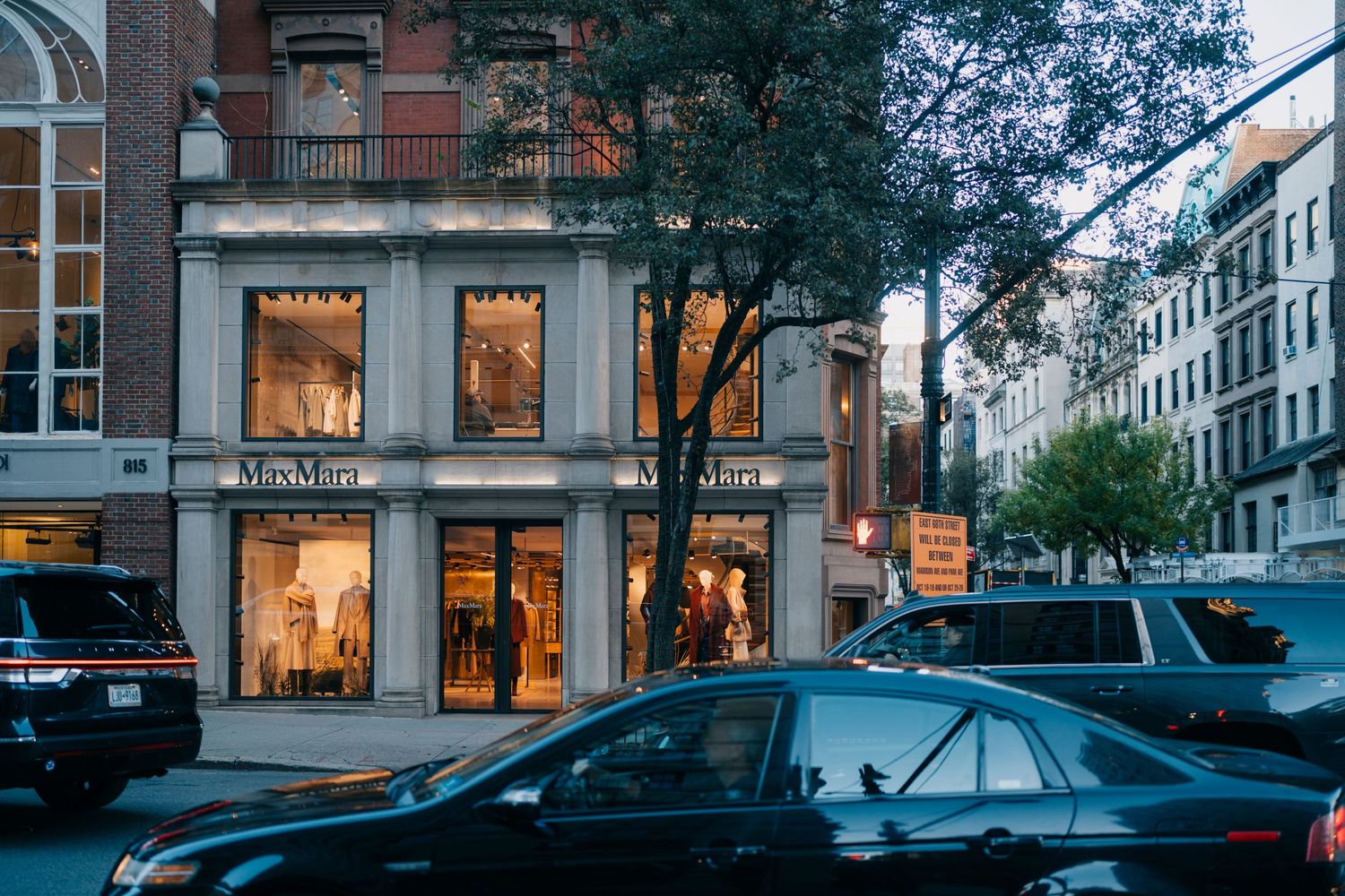 Urban street scene at dusk with illuminated storefronts, parked cars, and trees lining a European-style building facade.