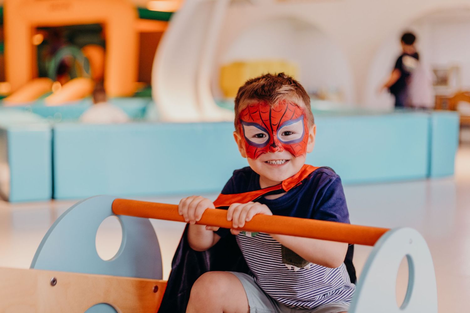 Child wearing Spider-Man face paint riding in play vehicle.