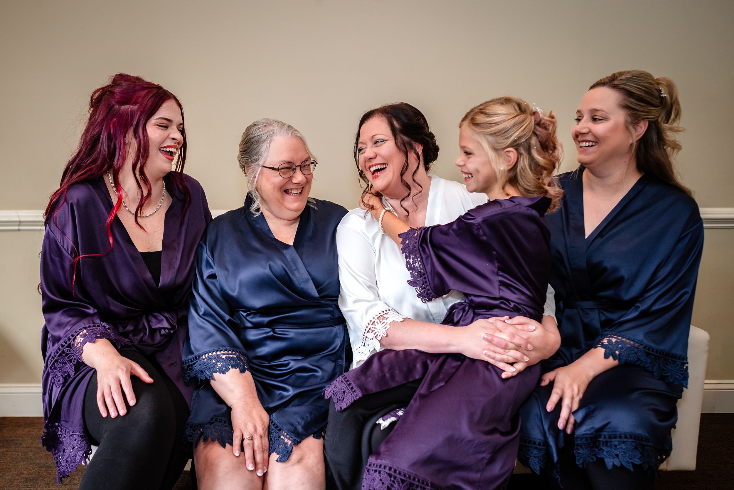 Wedding party members in satin robes sit together sharing a joyful moment before the ceremony.