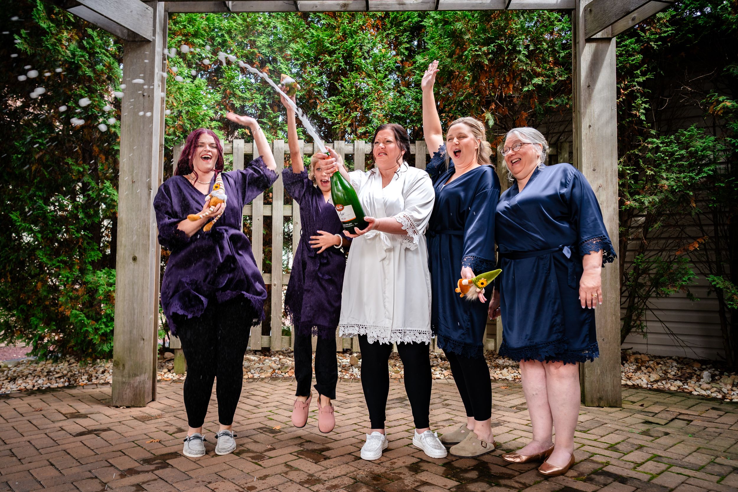 Wedding party poses under an outdoor pergola surrounded by lush greenery.