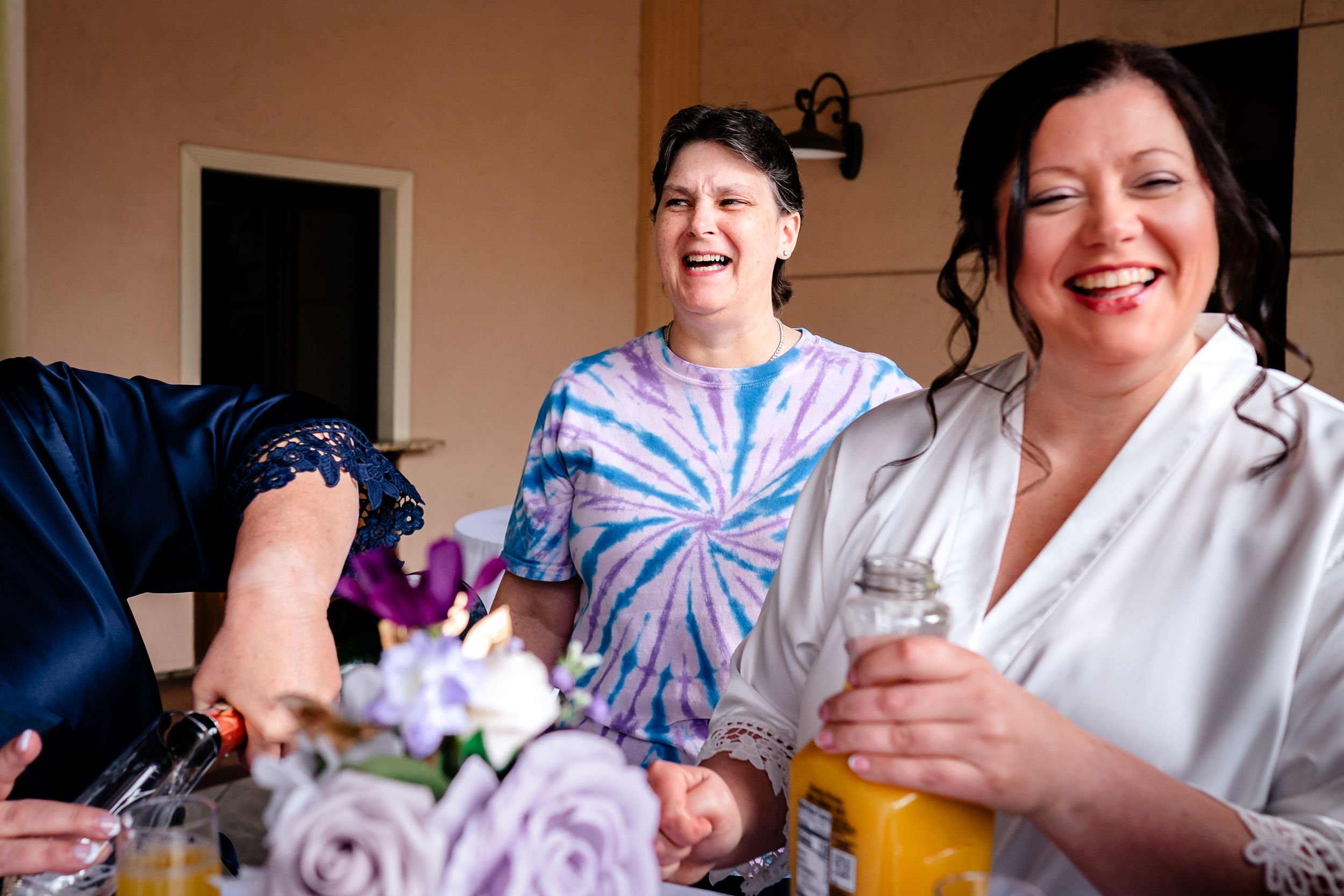 Two people share laughter and drinks during wedding preparations in a casual indoor setting.