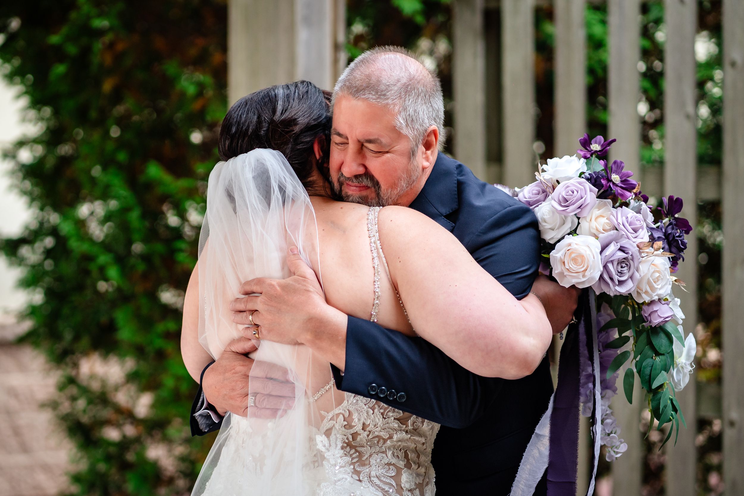 Intimate wedding ceremony moment under a white lattice pergola with purple floral bouquet.