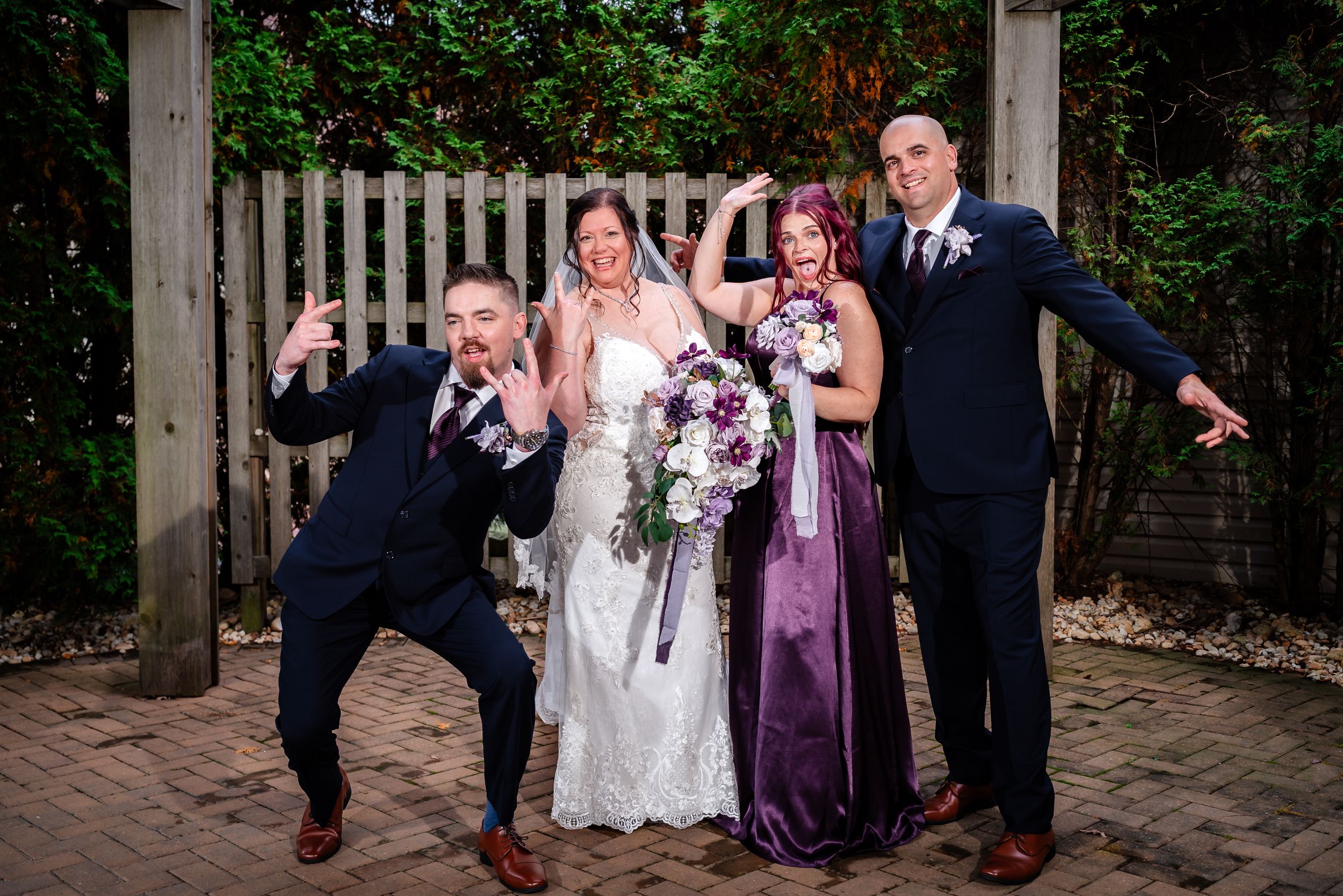 Wedding party poses together outdoors in purple and burgundy attire near a wooden fence.