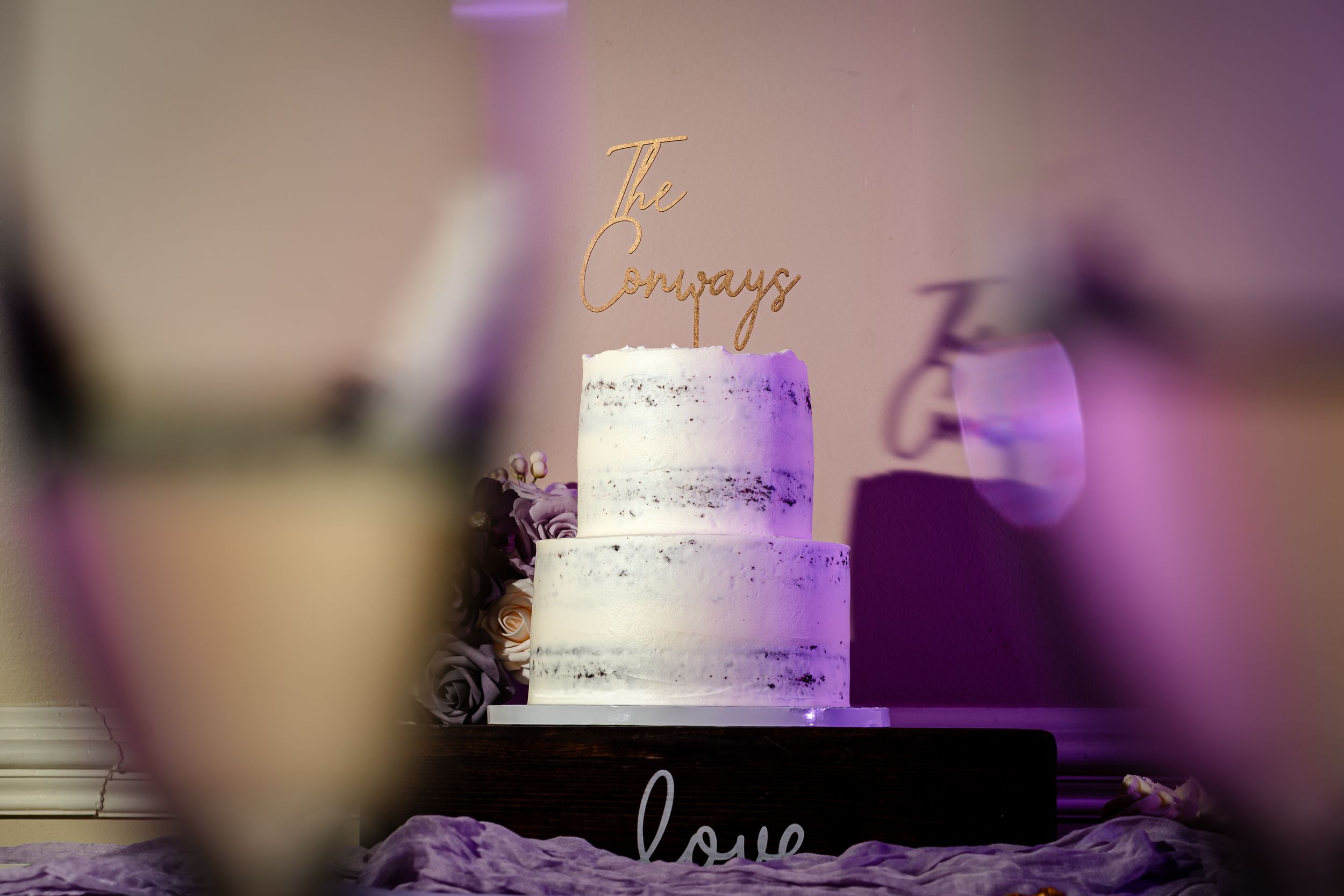 Elegant white wedding cake with purple lighting and decorative text.