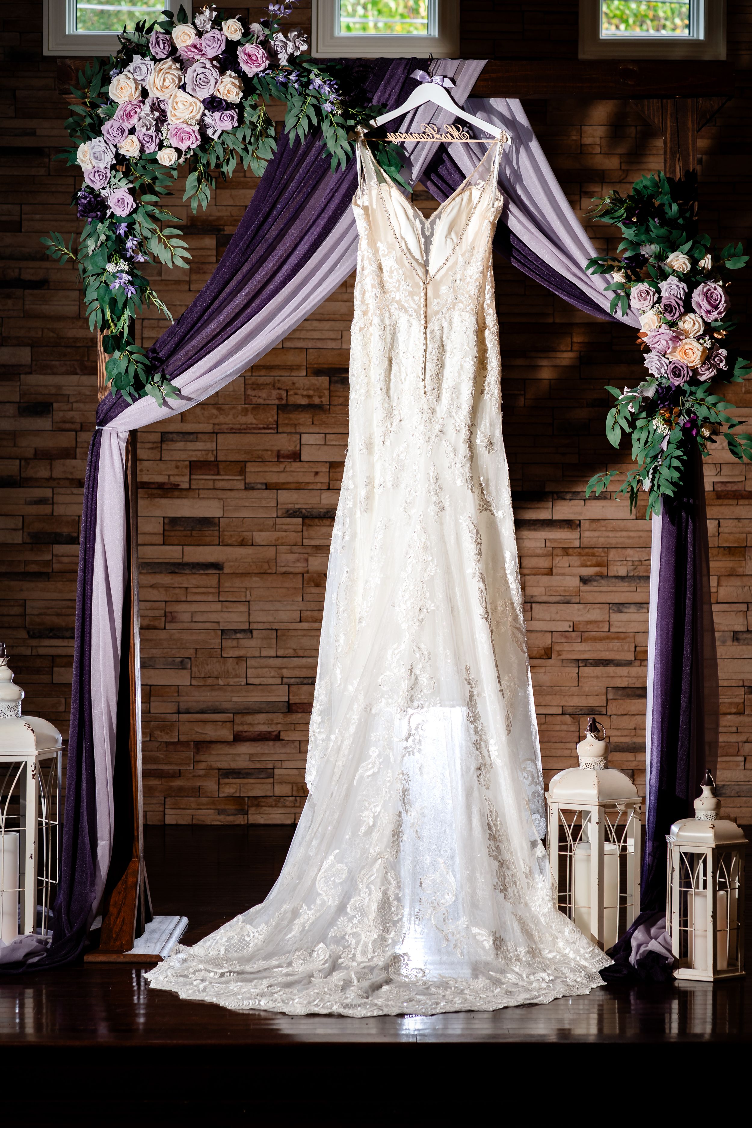 Elegant white wedding dress displayed against stone wall with purple draping and floral decorations.