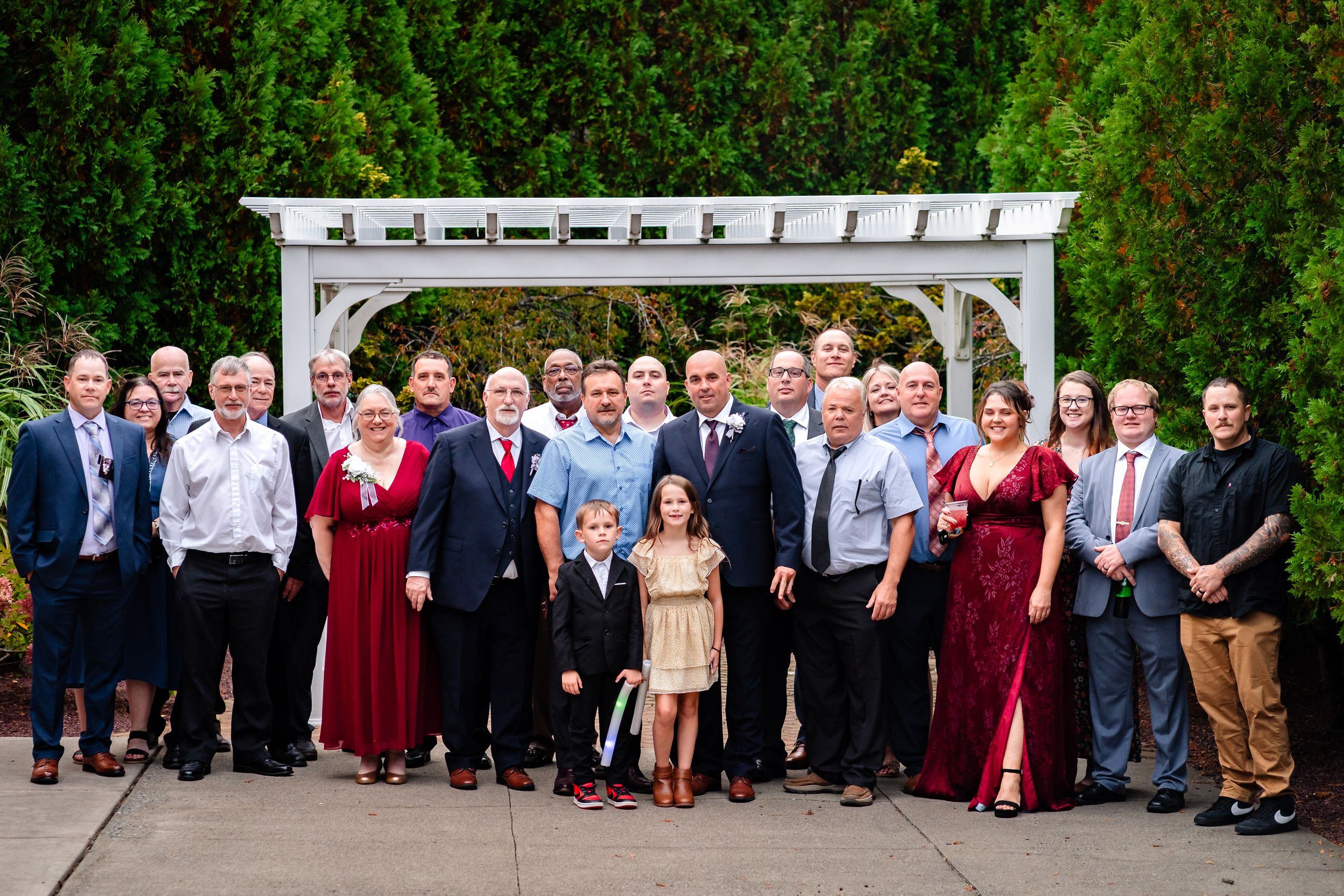 Large group wedding photo of guests and wedding party lined up outdoors under white pergola.