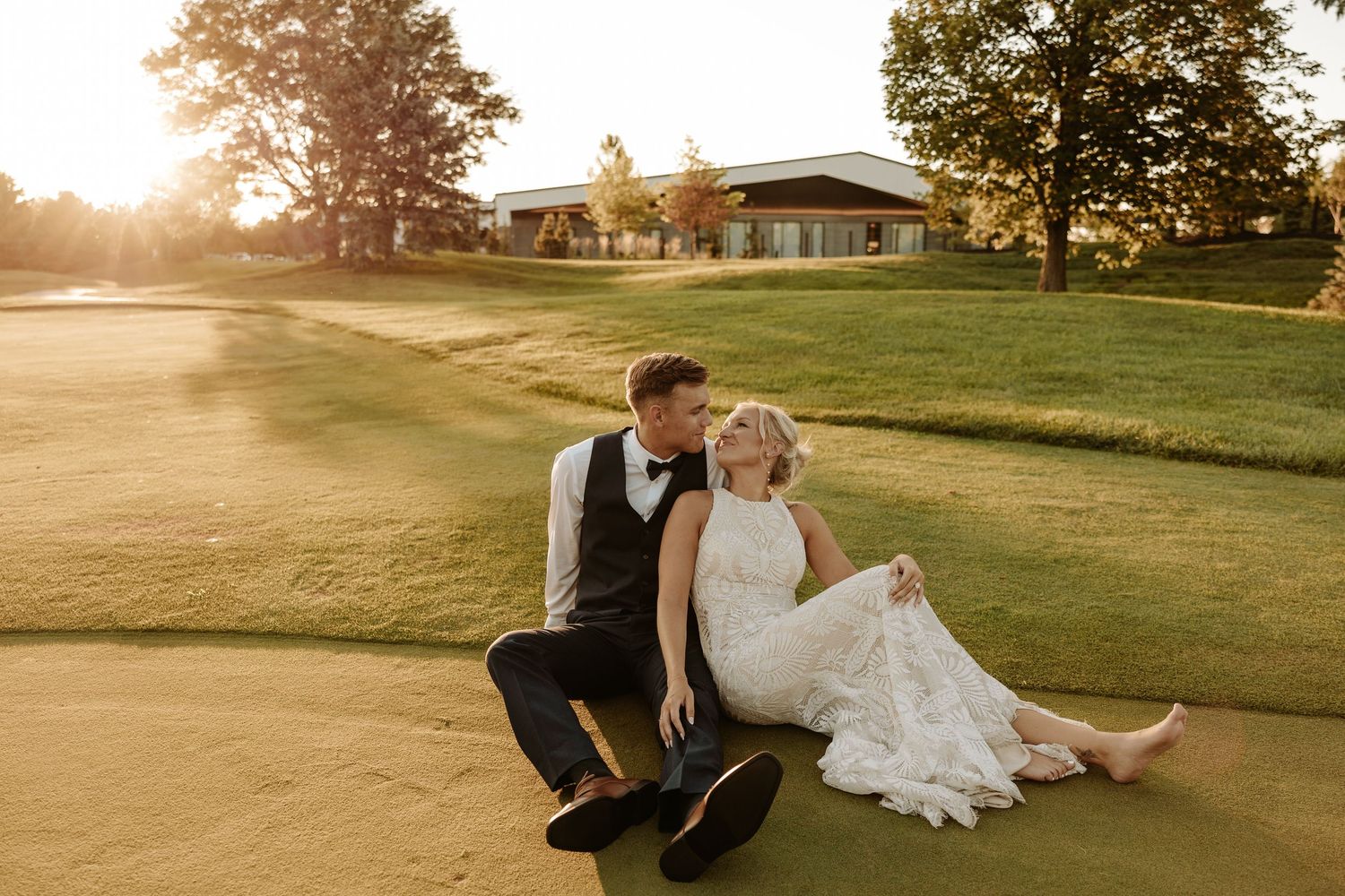 A couple in wedding attire sits together on a lush green golf course at sunset.