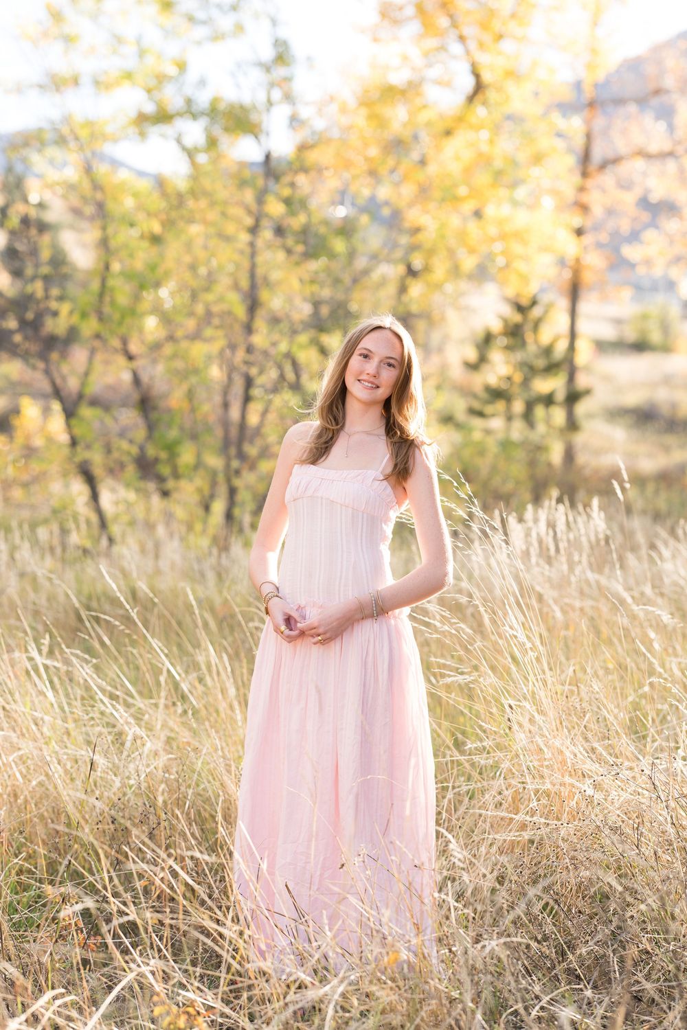 A woman in a flowing pink maxi dress stands in a sunlit field of tall grass with autumn foliage in the background.