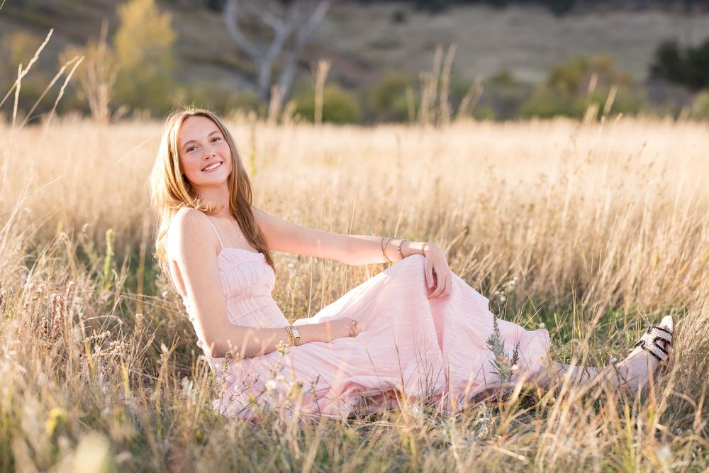 A person in a flowing pink dress poses gracefully in a sun-lit field of tall golden grass during sunset.