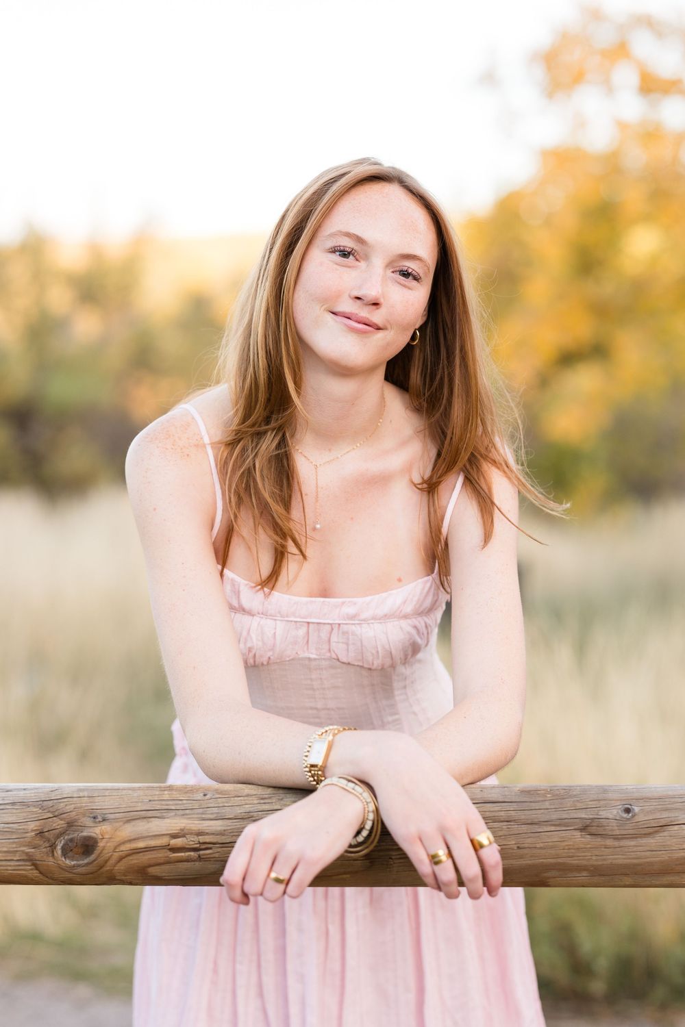 A portrait of someone in a pink dress leaning on a wooden fence with autumn foliage in the background.
