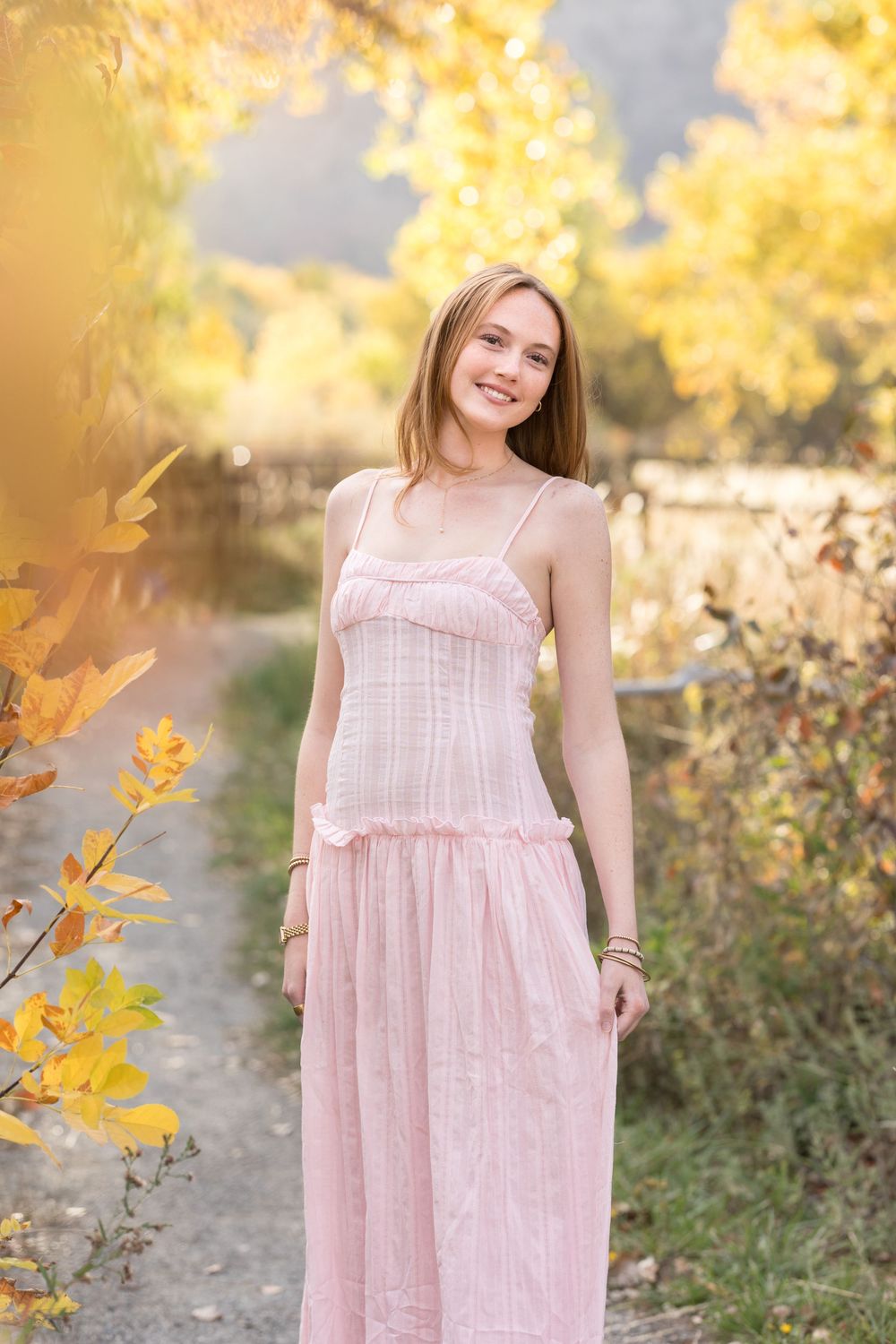 A soft pink babydoll dress with ruffle details is photographed against golden autumn foliage in natural lighting.