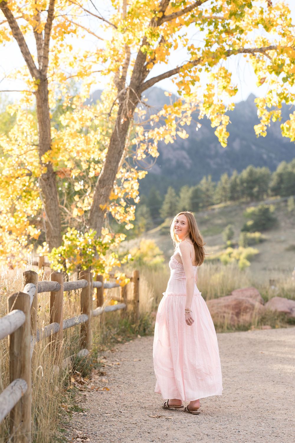 A pink dress flows gracefully along a rustic fence-lined path with golden autumn trees in the background.