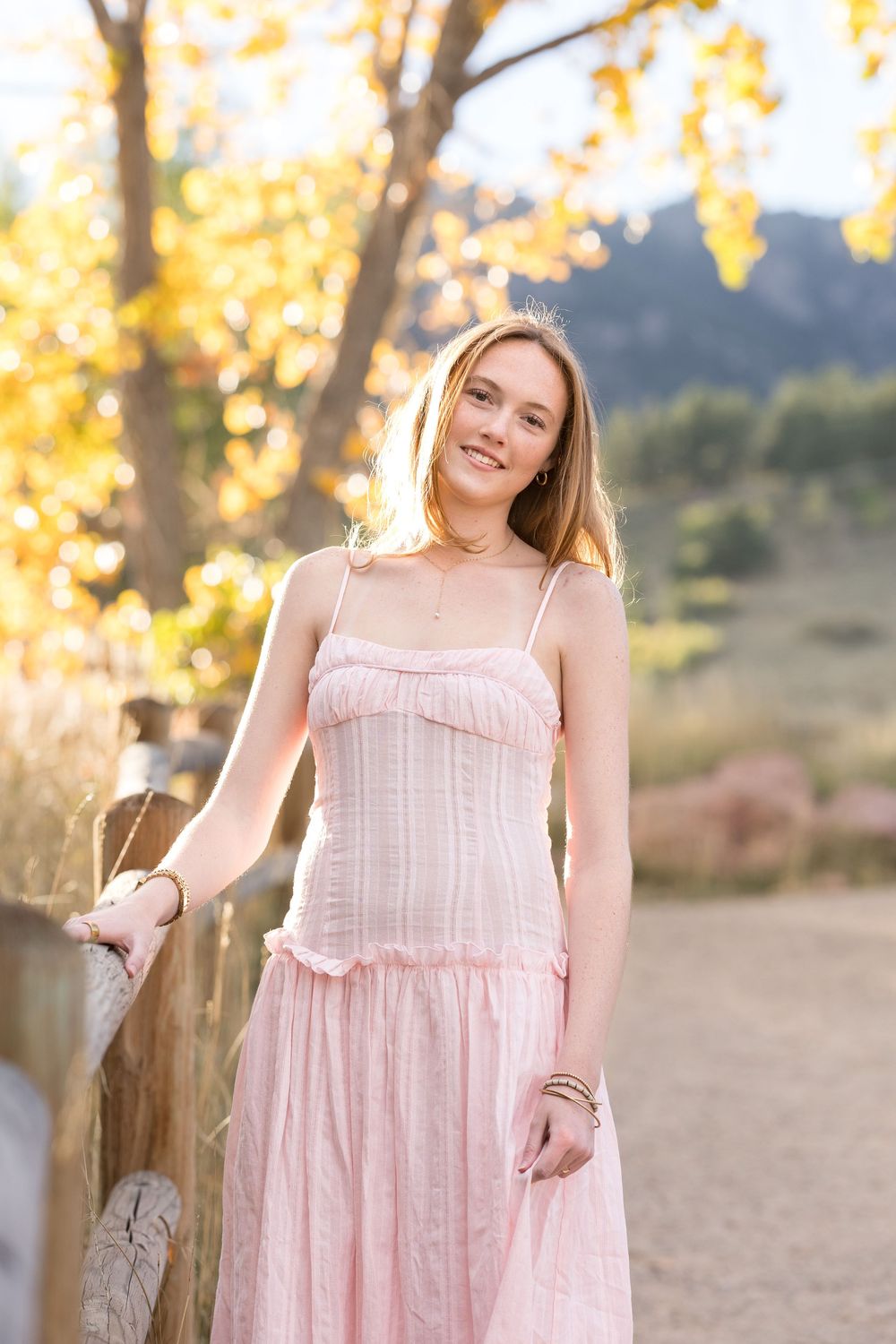 A feminine pink sundress with gathered tiers and pockets is showcased in a warm autumn outdoor setting.