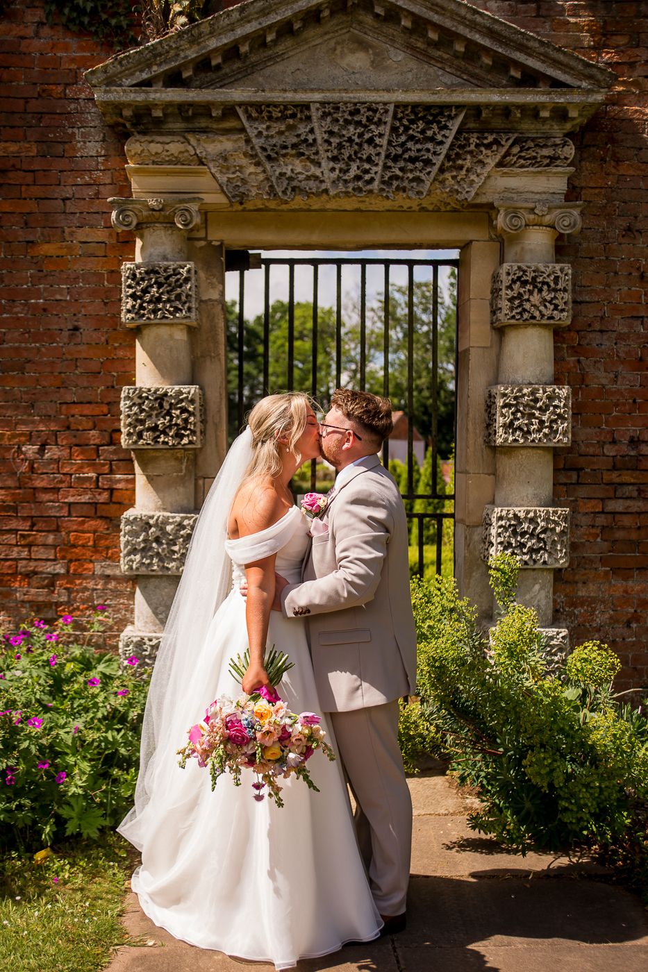 Wedding couple poses in garden archway with ornate stone columns and blooming flowers.