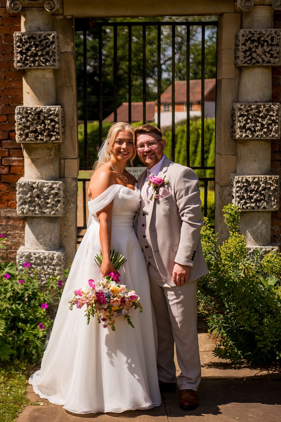 Newlyweds share intimate moment under ornate stone colonnade with flowering garden.