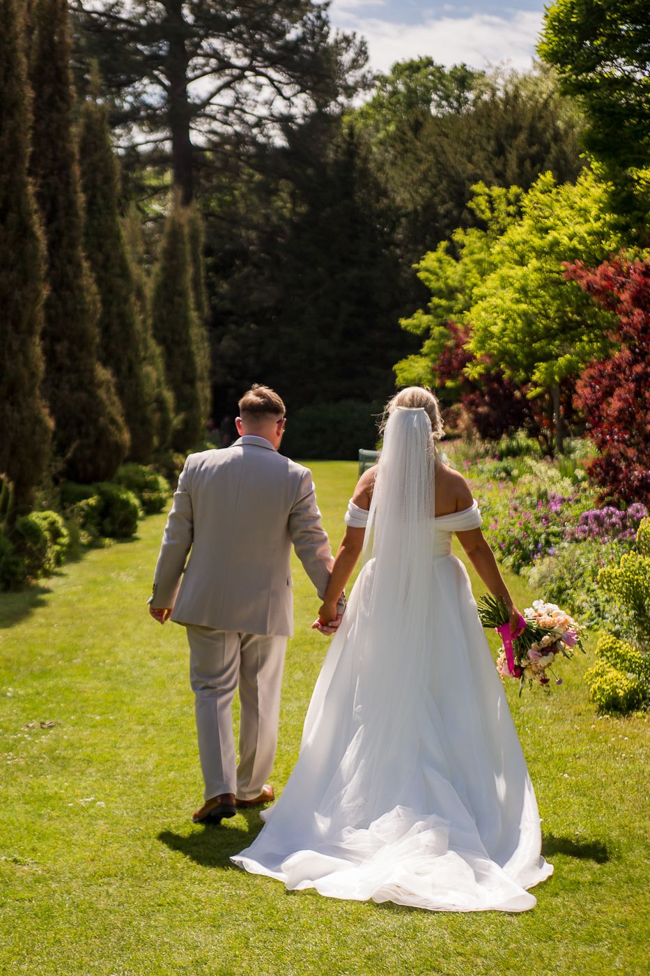 Wedding couple walks through lush garden pathway on sunny afternoon.