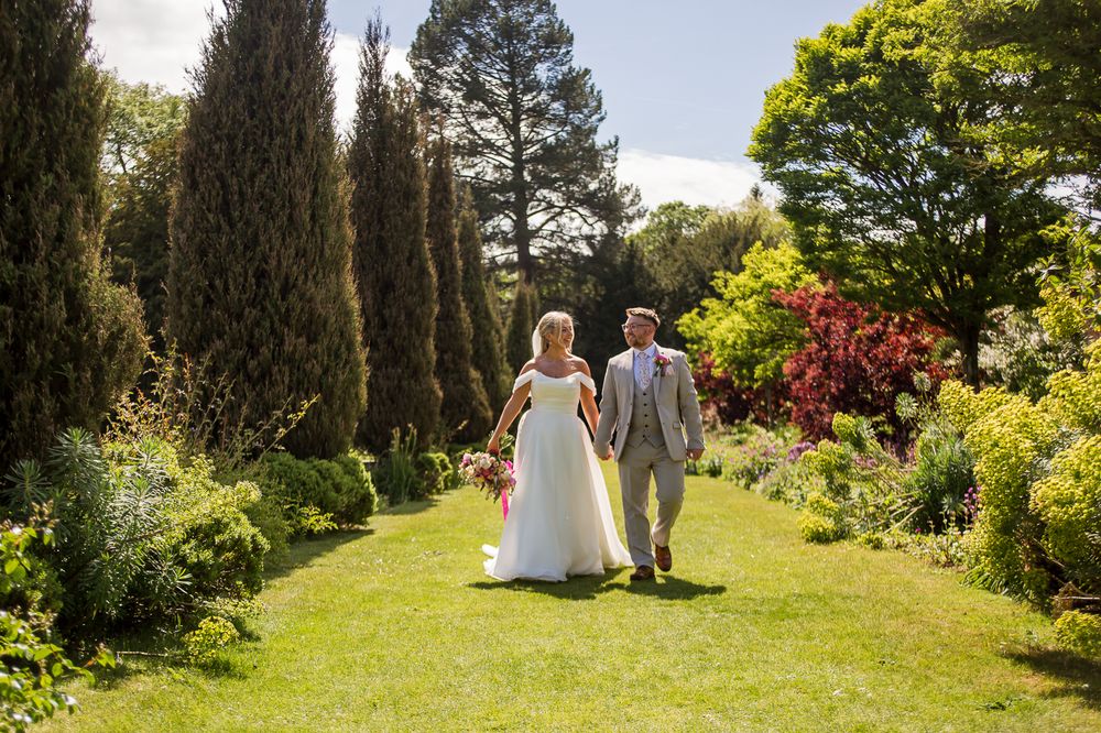 A couple walks through a lush garden path on their wedding day surrounded by greenery and flowers.