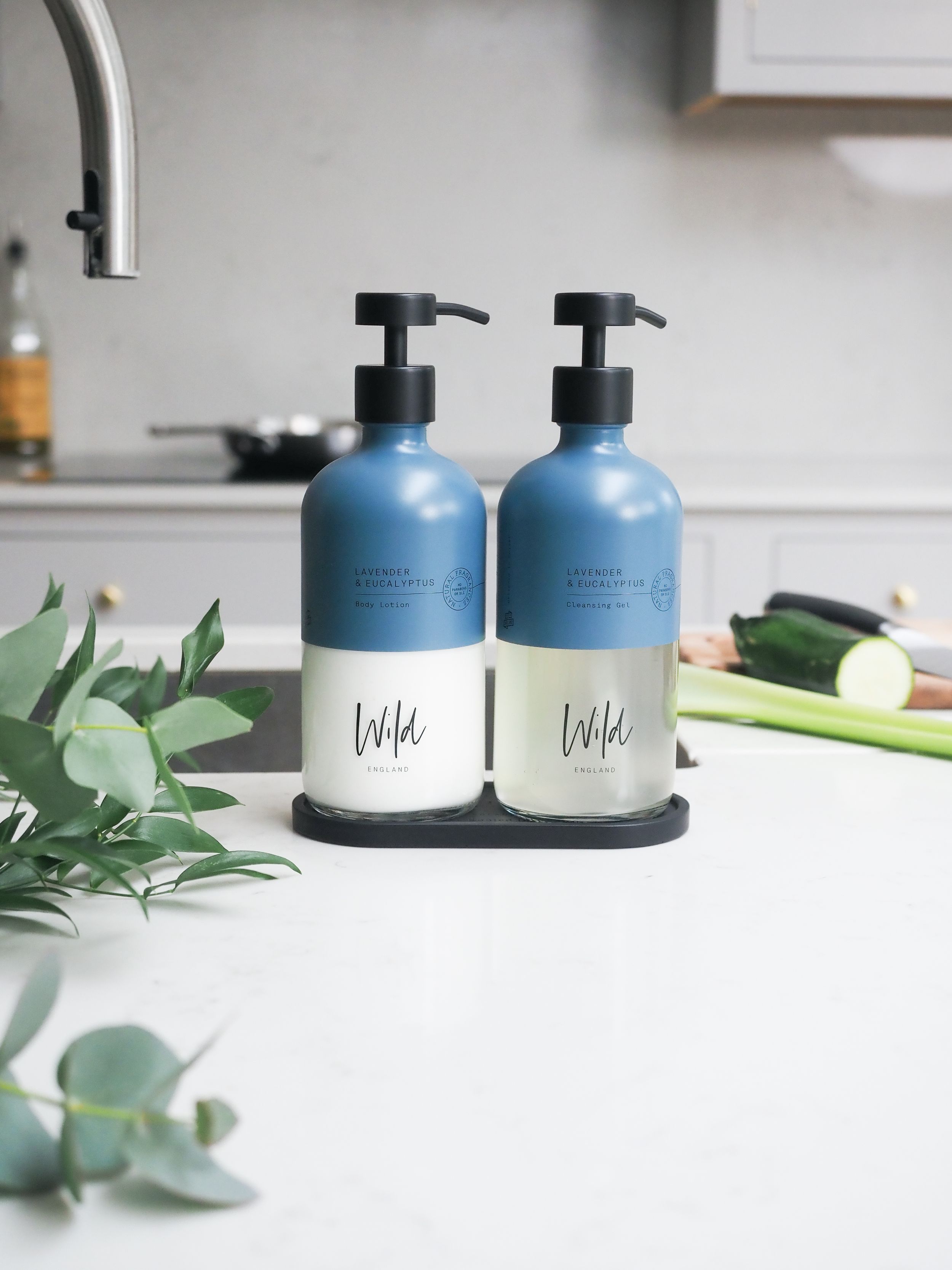 Two blue and white pump dispenser bottles on a black tray beside eucalyptus on a kitchen counter.