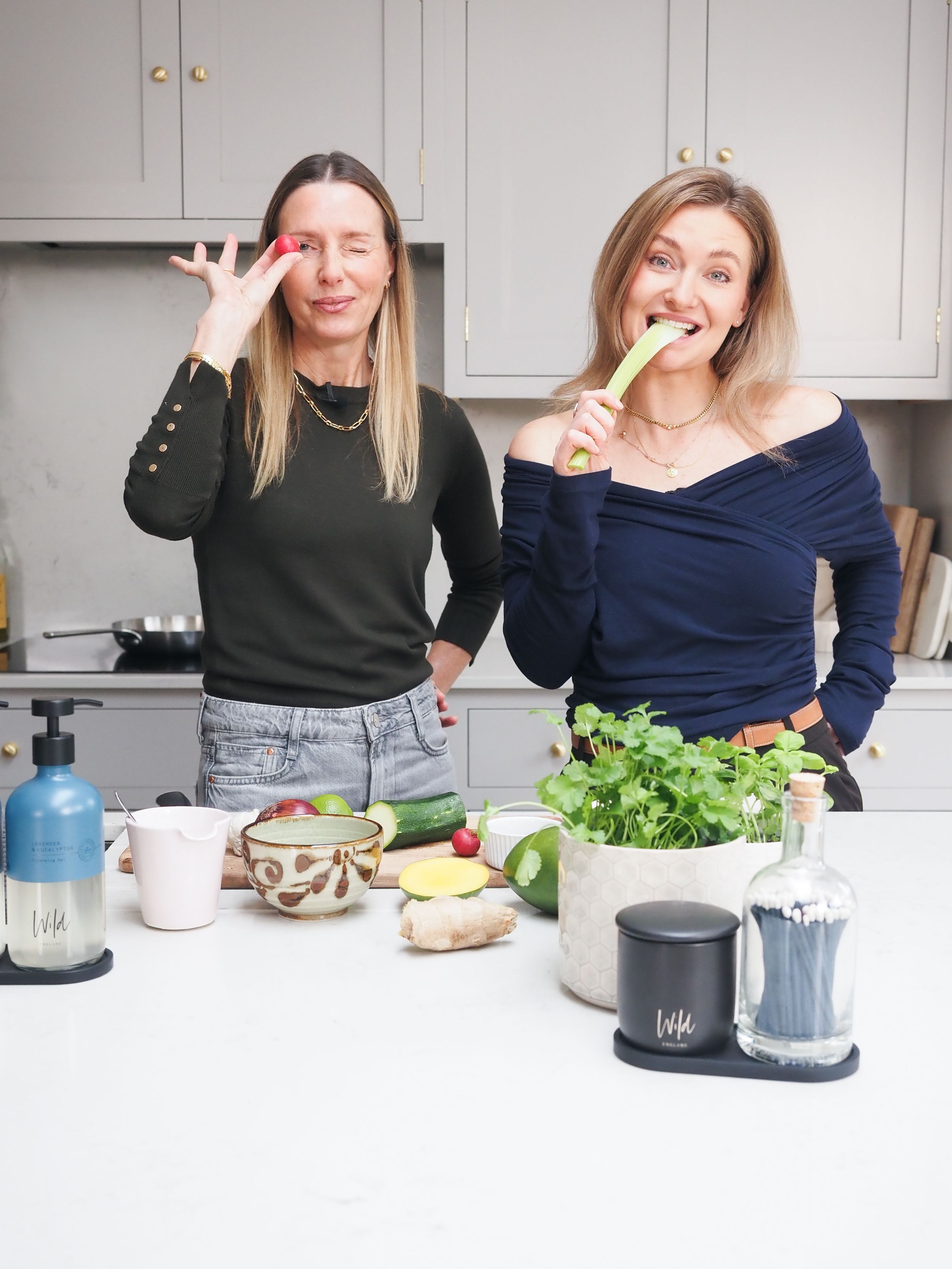 Two women in a modern kitchen playfully posing with fresh vegetables including celery and lime on a white counter.