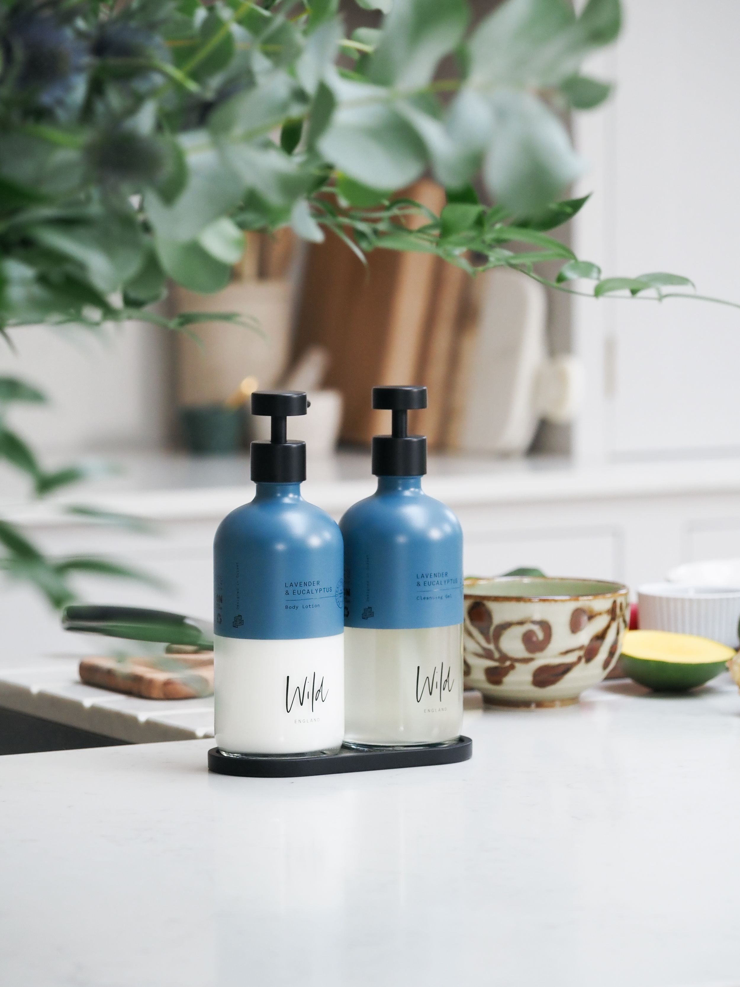 Two blue pump bottles on a black tray sit on a white kitchen counter beside a ceramic bowl and green eucalyptus.