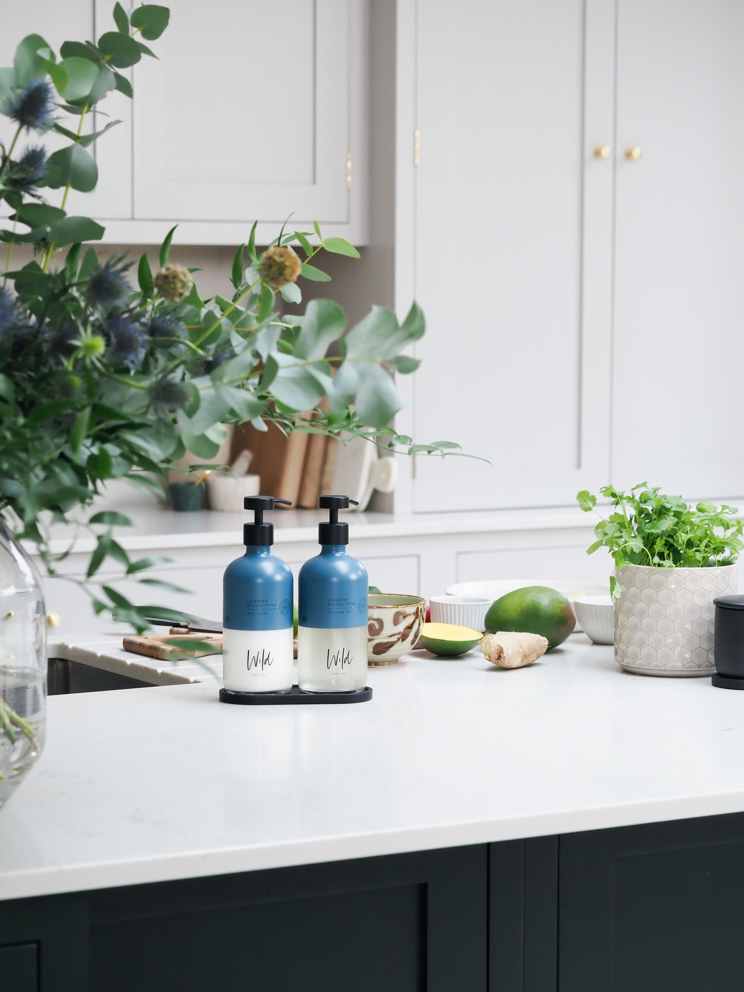 Two blue ceramic soap dispensers on a white kitchen counter with fresh herbs, lime, and green plants in background.