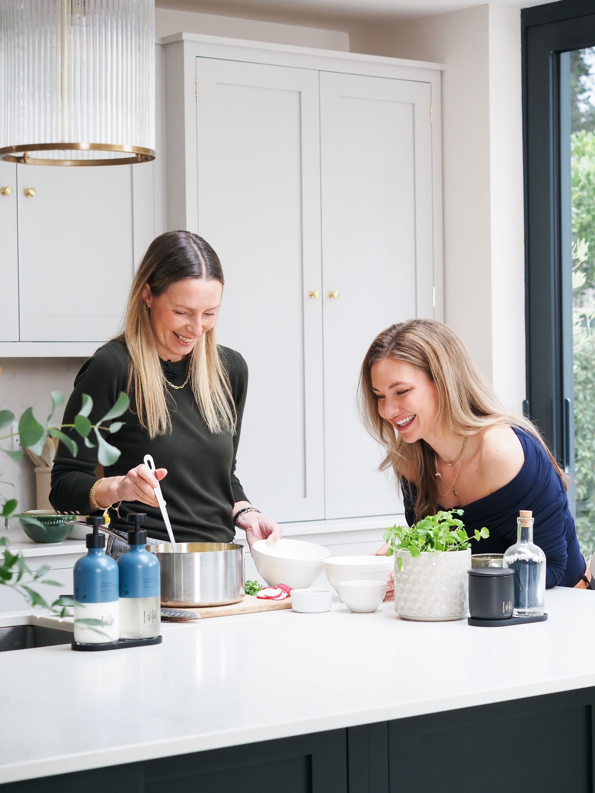 Two women laughing and cooking together in a stylish modern kitchen with green cabinetry.
