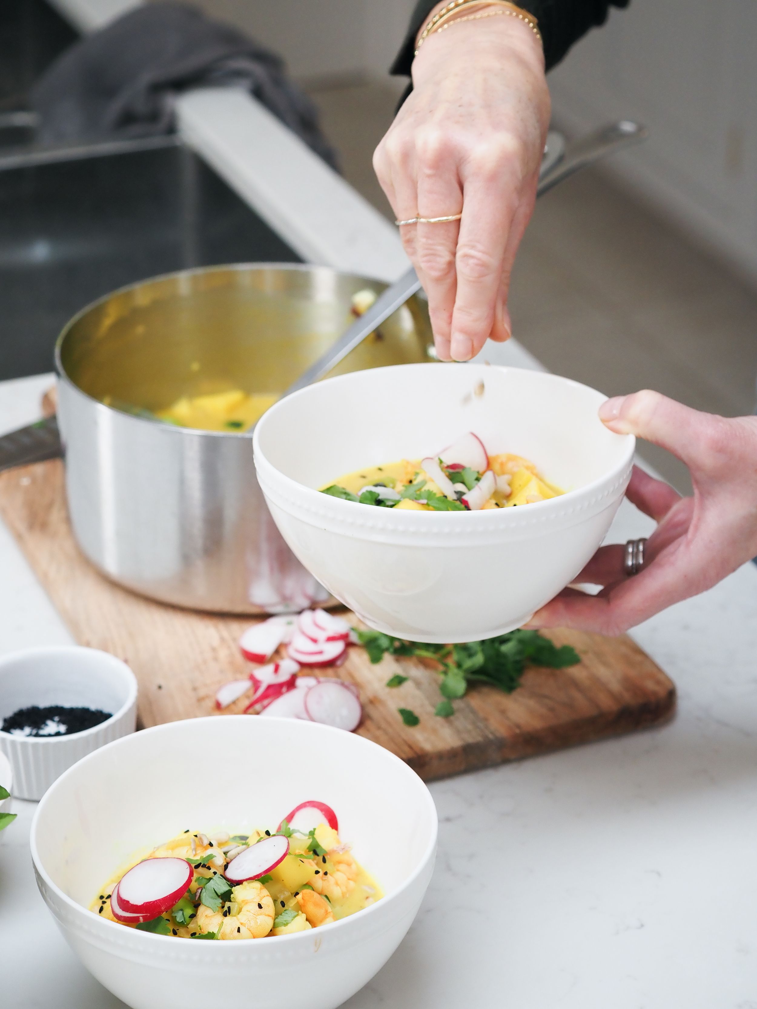 Hands serving a colorful vegetable soup into white bowls on a wooden cutting board with fresh garnishes.