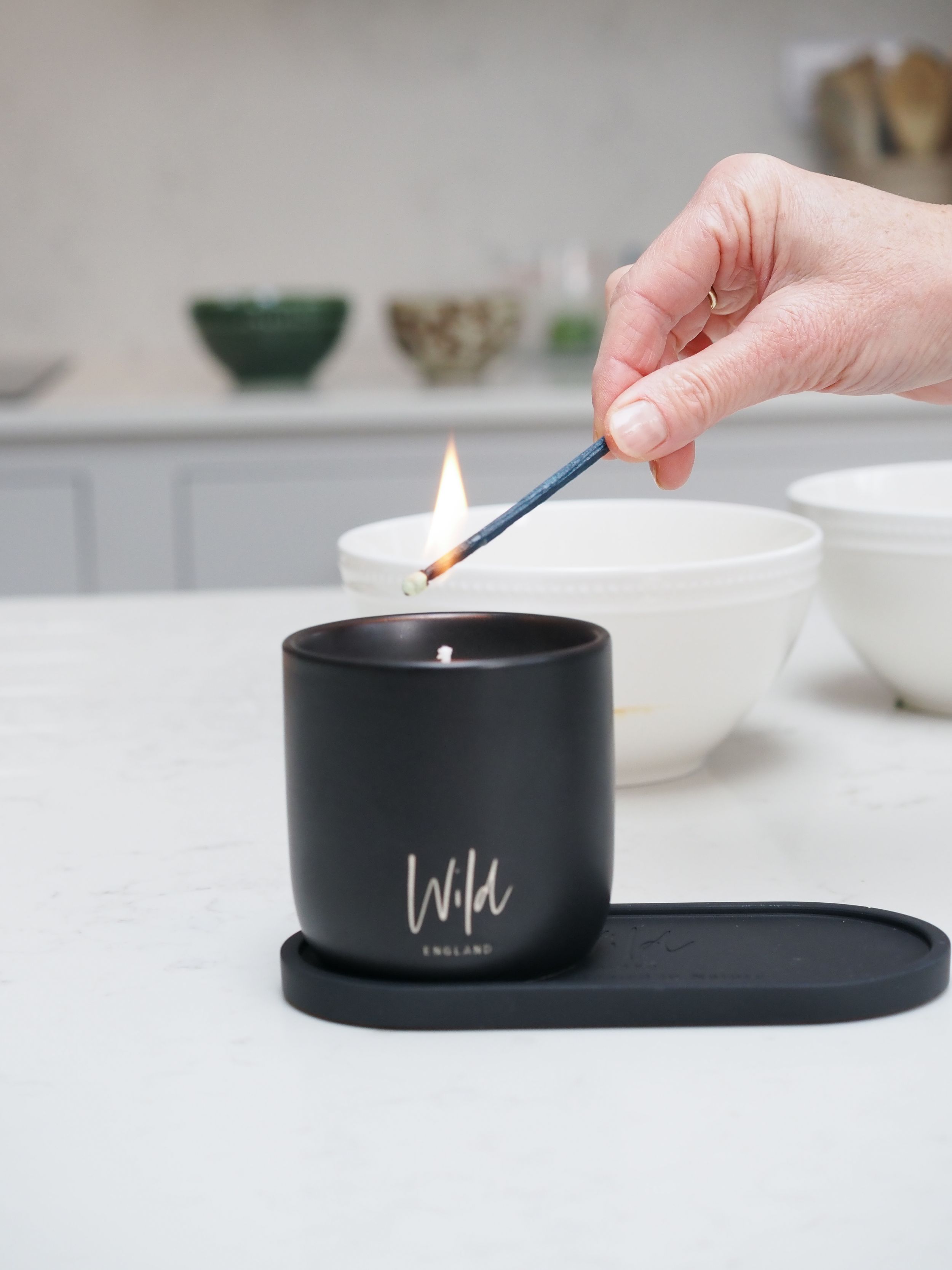 Hand lighting a black Wild England candle on a white marble kitchen countertop with a match.