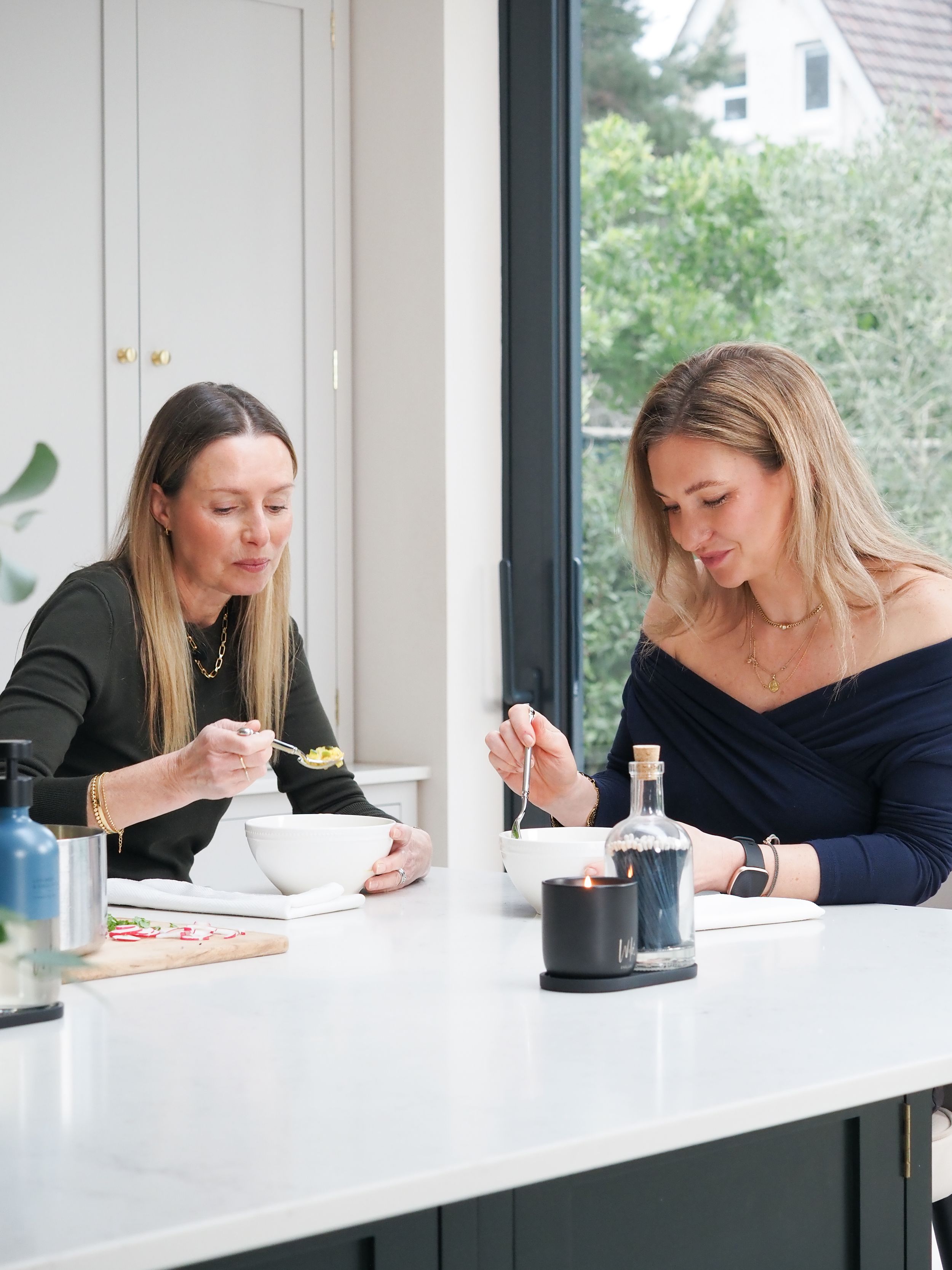 Two women sitting at a white kitchen counter eating from bowls with spoons in a modern bright kitchen.