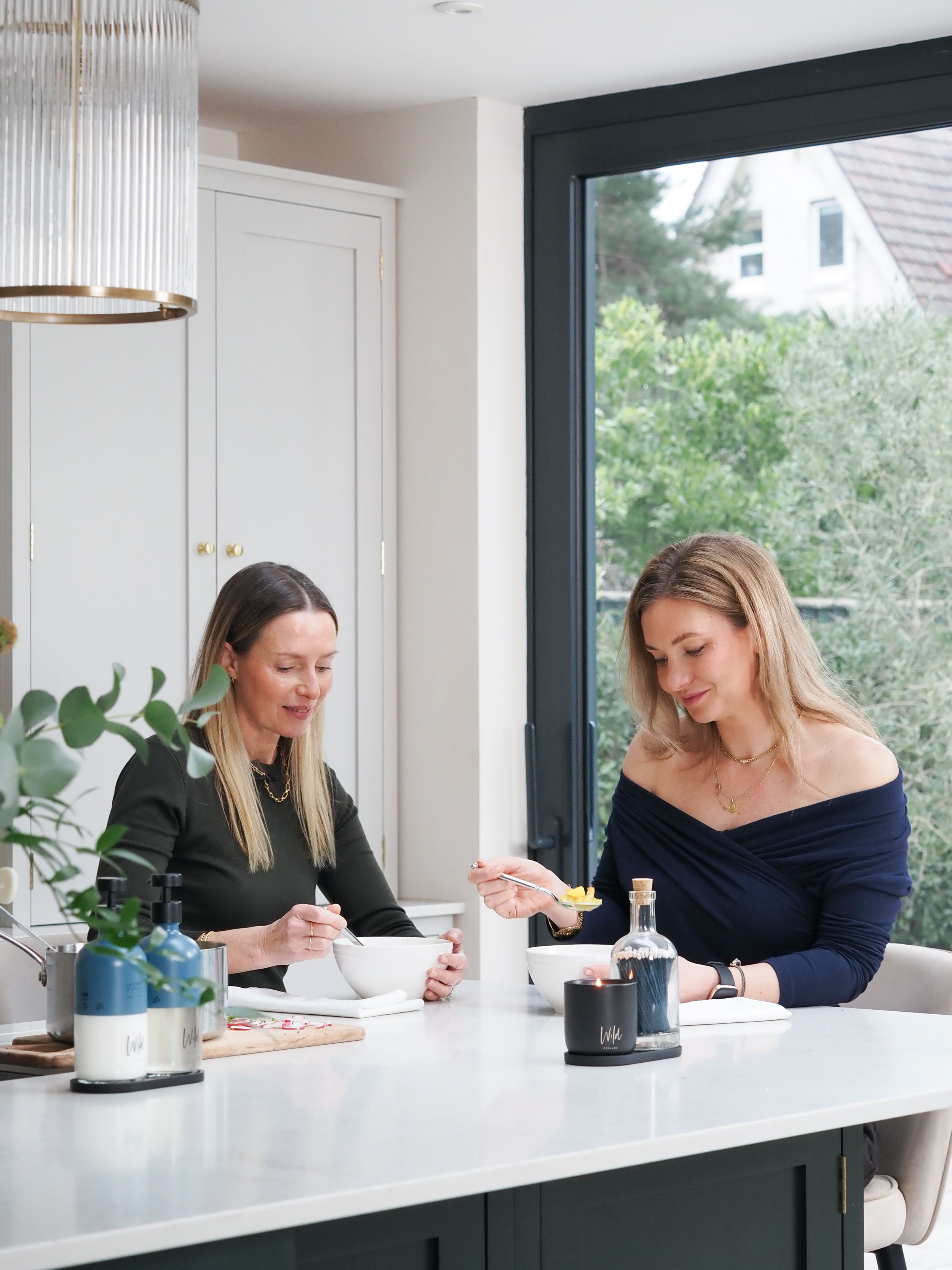 Two women enjoying a meal together at a modern kitchen island with green cabinetry and large windows.