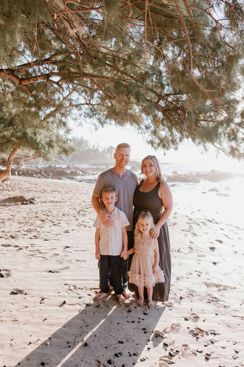 A family stands together on a sunny beach under pine tree branches casting shadows on the sand.
