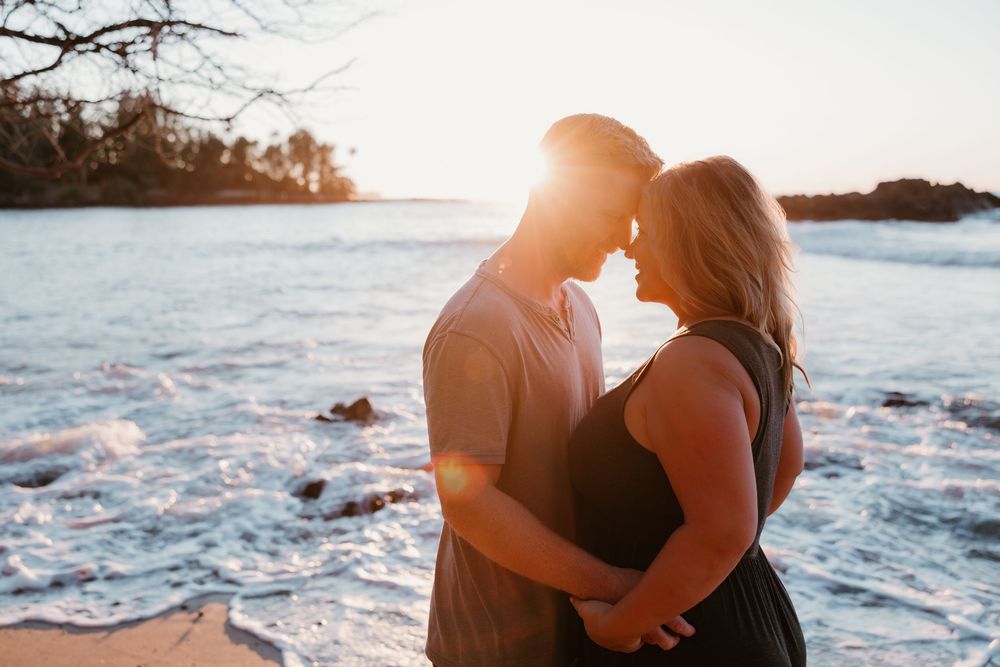 Romantic couple embraces on rocky beach at sunset with ocean waves crashing in background.