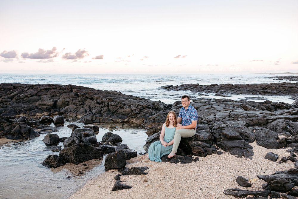 A couple sits together on volcanic rocks at the beach during sunset in Hawaii.