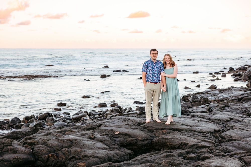 Two people stand on black lava rocks at sunset along a Hawaiian beach with waves crashing in the background.