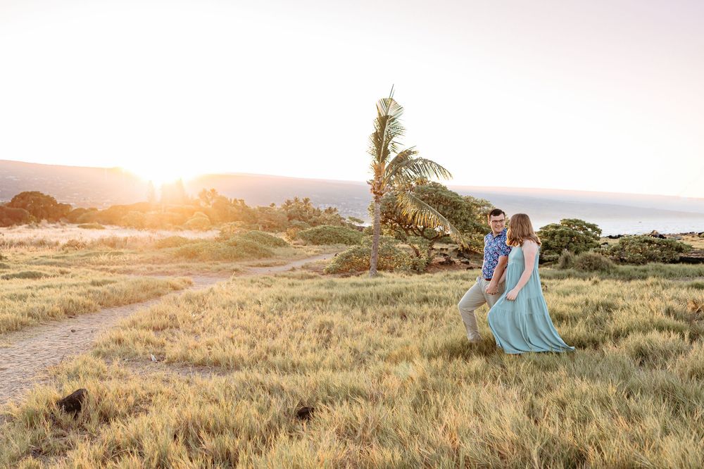 Romantic couple embraces in golden sunlit field with palm trees and ocean views at sunset in Hawaii.