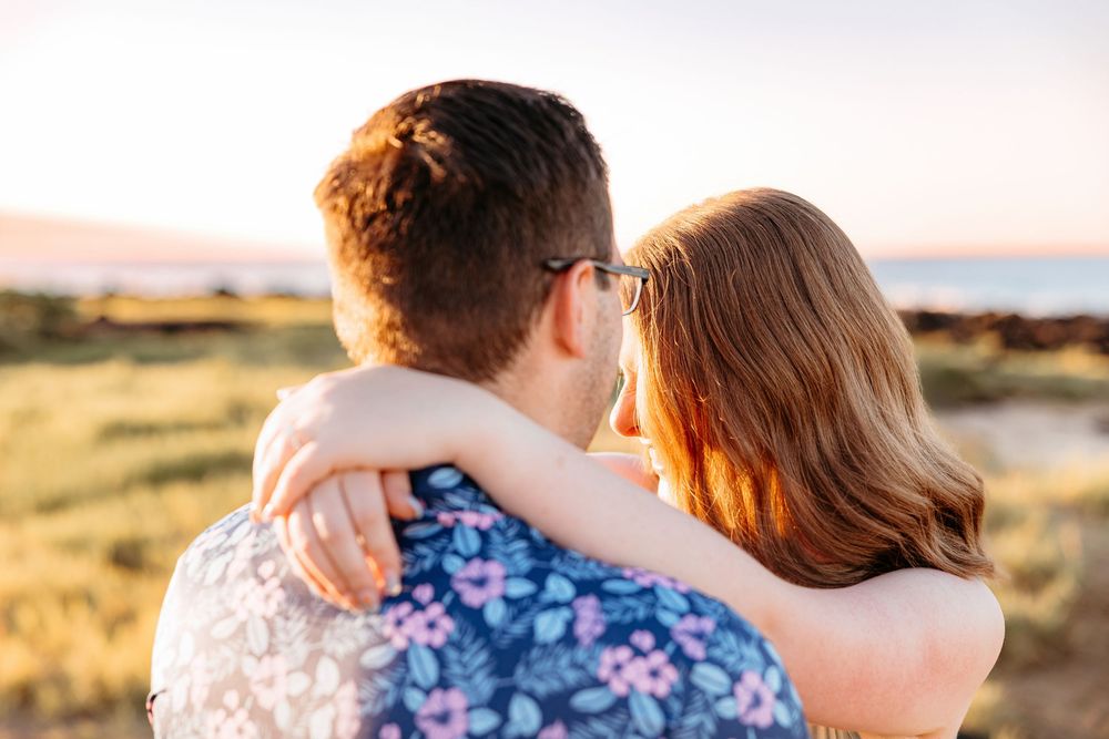 A couple embraces while watching a golden sunset over grassy hills near the water.