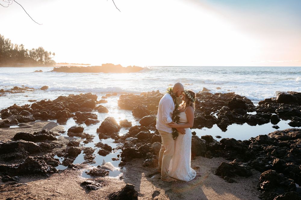 A couple stands on a rocky beach at sunset with waves crashing against the shore in the background.